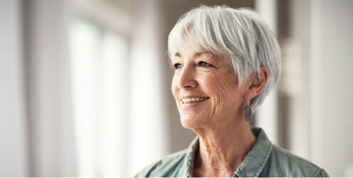 Smiling older woman with short silver hair, wearing earrings and a casual top, looking off to the side in a bright indoor setting.