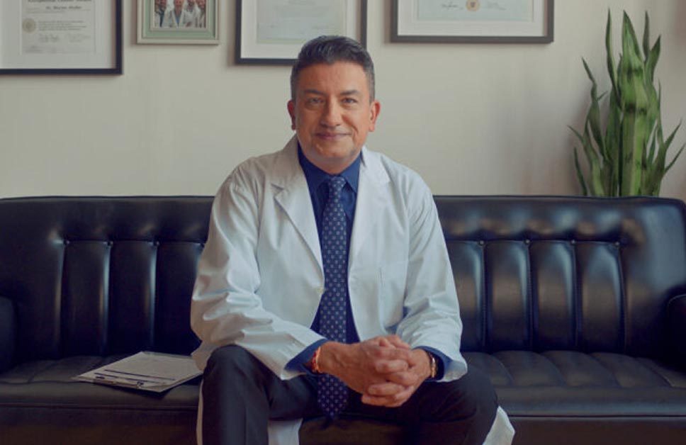 Clinical investigator in a white lab coat sitting on a black leather couch, with framed medical certificates in the background.