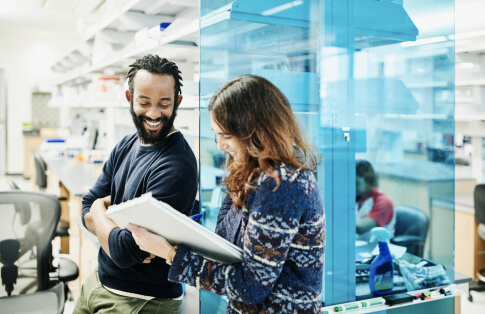 Two smiling researchers discussing notes in a bright laboratory, with scientific equipment and workstations in the background.